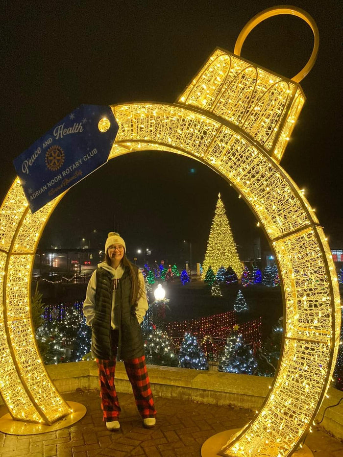 A woman bundled up in winter clothes smiles under a lighted walk-through ornament with a giant Christmas tree in the background.