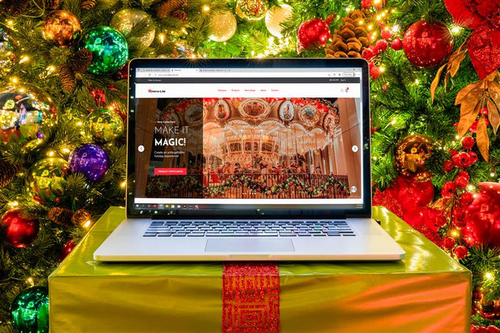 A laptop sits on top of a gift box in front of a Christmas tree. The laptop screen shows the dekra-lite.com homepage.