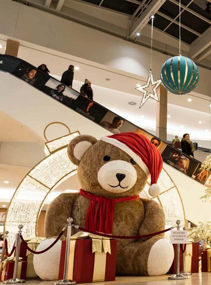 A shopping mall Christmas display with a giant inflatable teddy bear in front of a gold lit walk-through ornament. Burgundy gift boxes with gold bows surround the display. Christmas ornaments and stars hang in the distance.
