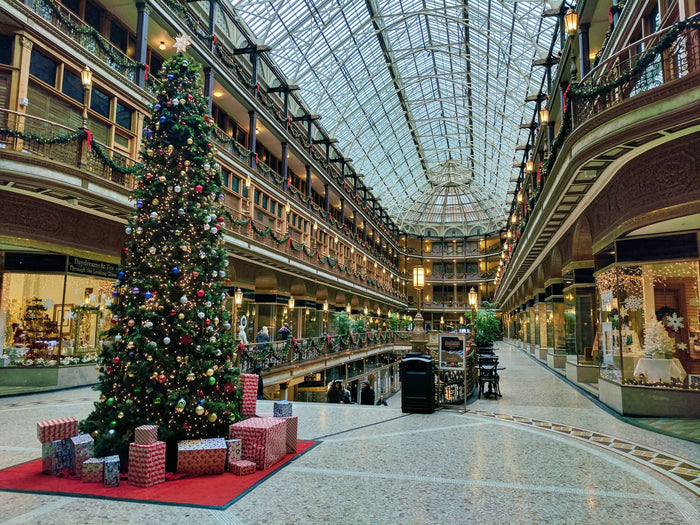 Large indoor shopping center decorated for Christmas with a giant Christmas tree on the left side of the image surrounded by gift boxes.