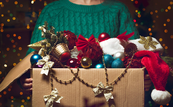 A person wearing a green sweater holds a box overflowing with Christmas decorations.
