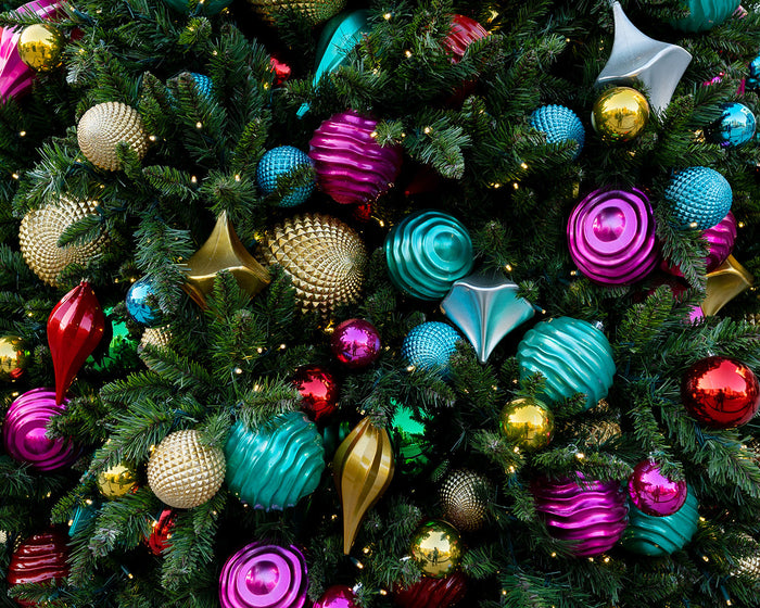 A close-up of a commercial Christmas tree decorated with teal, hot pink, gold, red, and green ornaments of various shapes, styles, and textures.