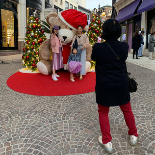 Two women and a child pose in front of a giant teddy bear Christmas photo op with two outdoor Christmas trees