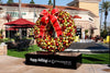 A giant wreath decorated with gold and burgundy Christmas ornaments and a red bow with gold trim stands on a custom black base that says 