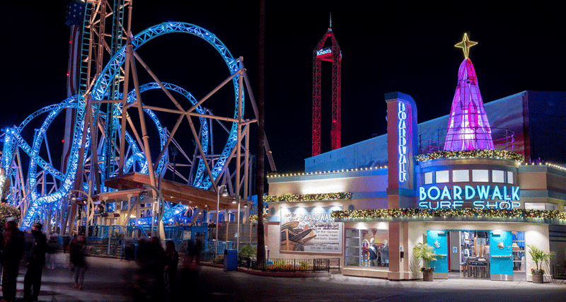 Amusement park storefront lined with foliage garland with a custom surfboard Christmas tree on the roof with a roller coaster in the background