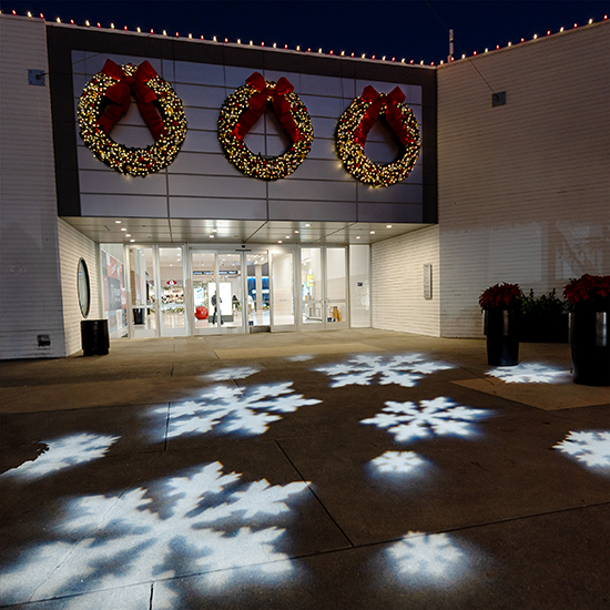 Projector displaying snowflakes on the ground in front of a department store containing 3 large lit wreaths