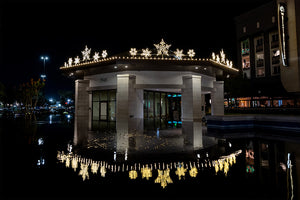 Lit snowflake decorations line the roof of a gazebo and are reflected in a fountain.