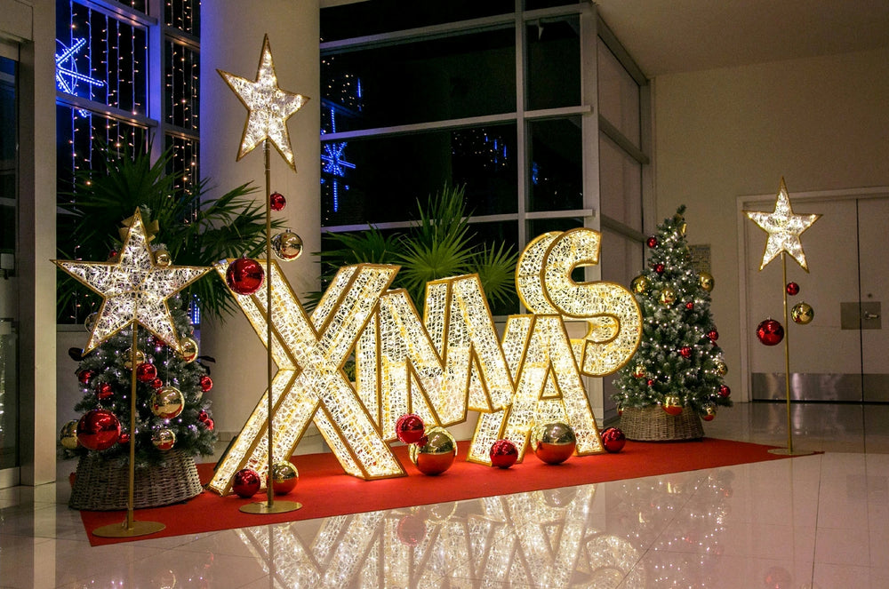 Giant Christmas prop letters that spell XMAS lit with warm white LED mini lights surrounded by oversized ornaments in red and gold, three giant gold glitter star props, and two commercial Christmas trees